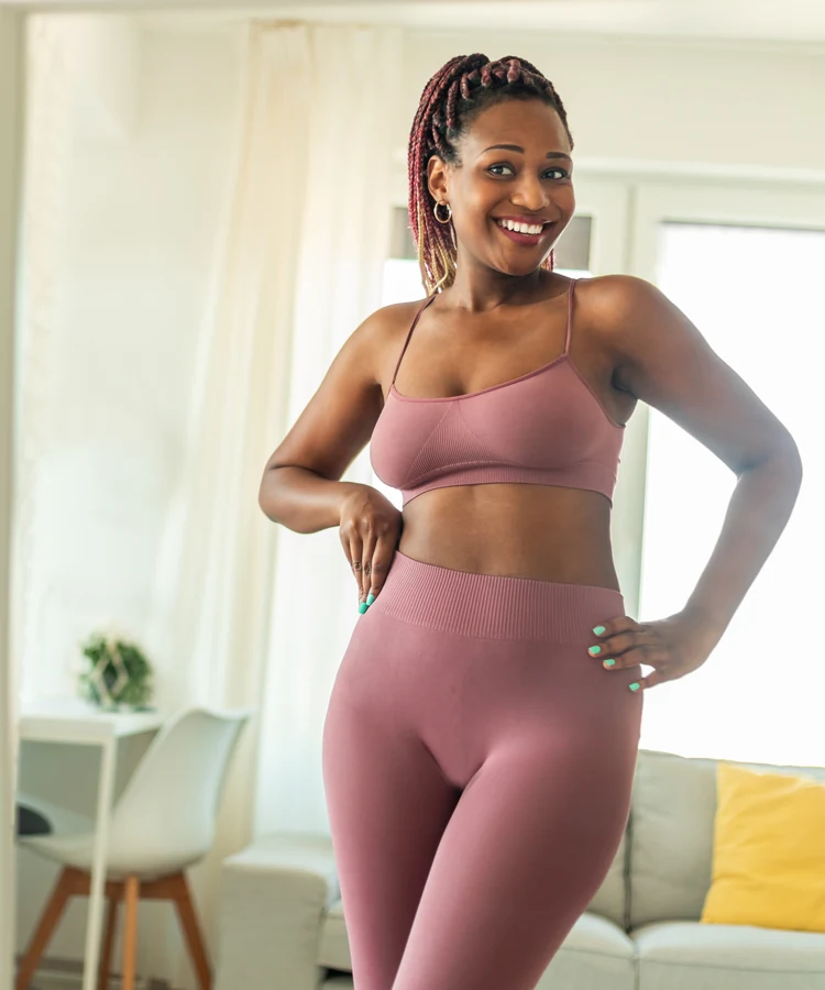 Confident woman in pink athletic wear standing in bright modern living room for fitness wellness