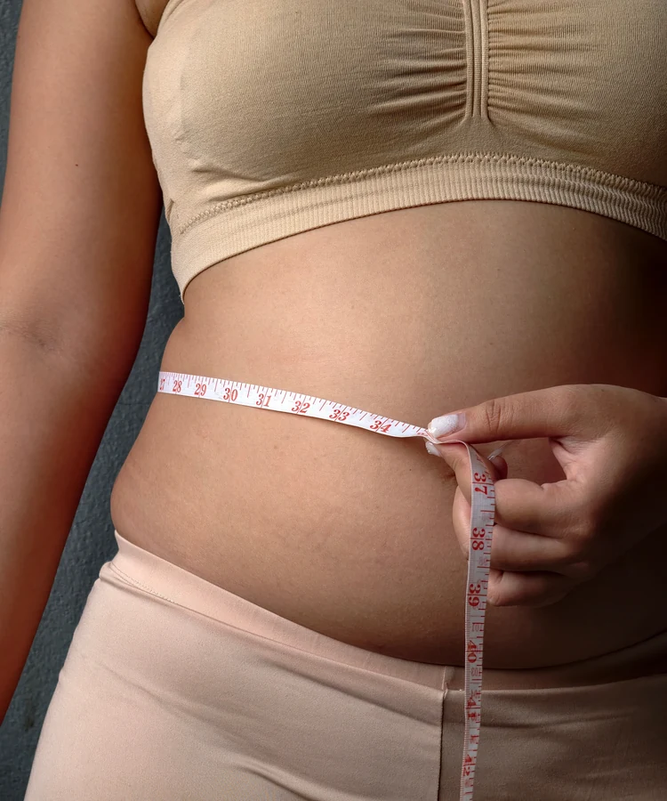 Pregnant woman measuring belly circumference with tape measure wearing beige activewear for prenatal wellness tracking