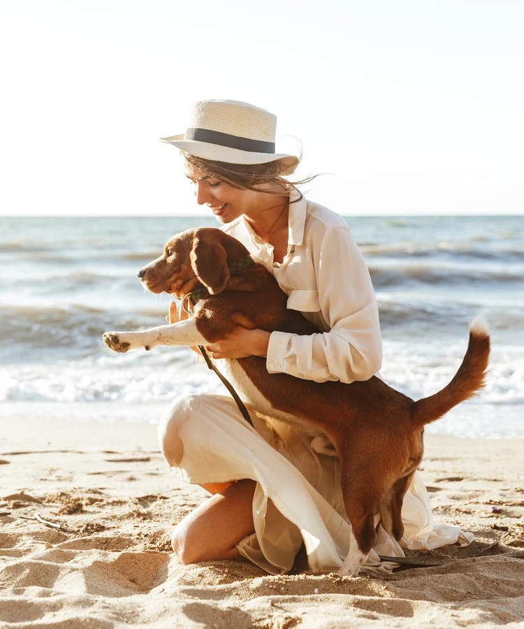 Woman in white hat enjoying peaceful moment with dog on sunny beach, wellness lifestyle concept