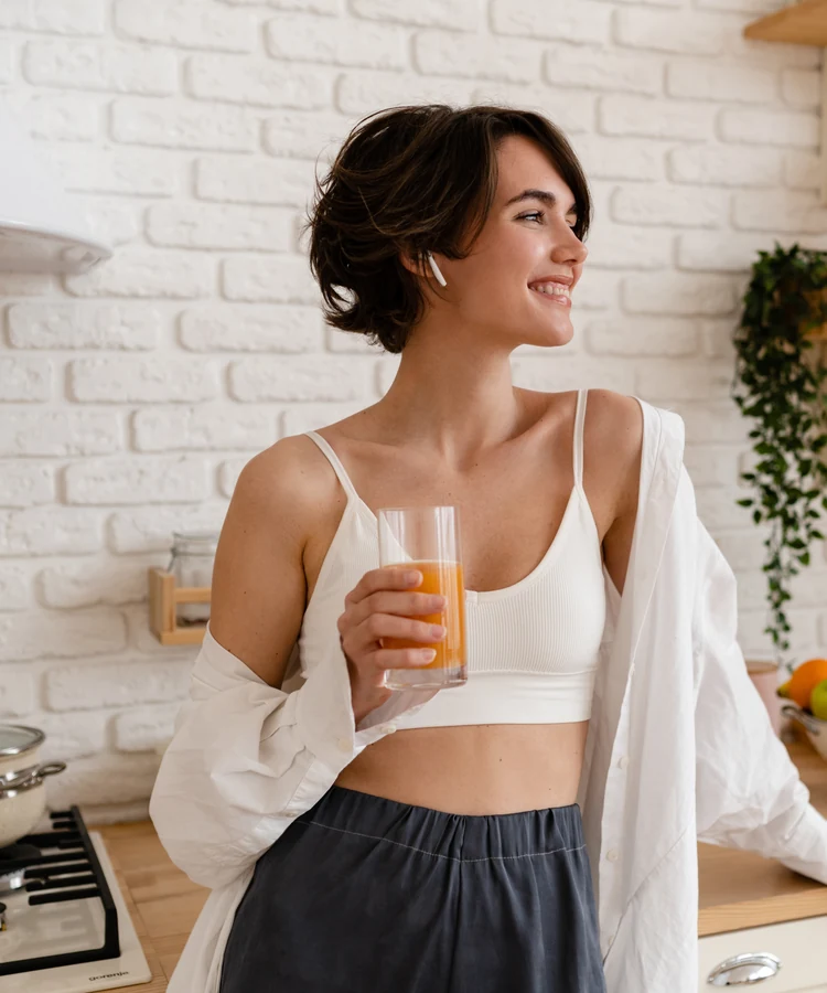 Woman in white sports bra drinking fresh orange juice in bright modern kitchen for healthy wellness lifestyle