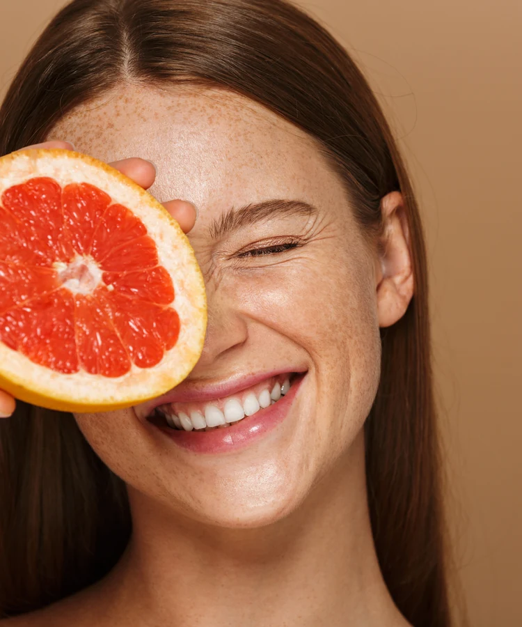 Happy woman with freckles holding fresh pink grapefruit slice over eye for natural wellness skincare
