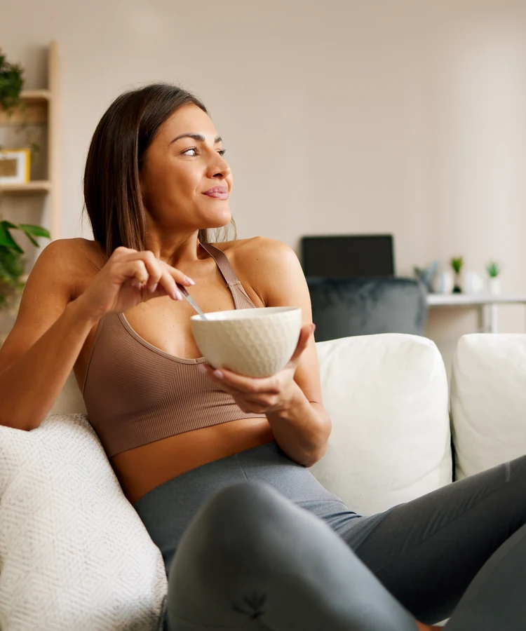 Woman in athletic wear enjoying healthy meal while relaxing on white couch in modern living room