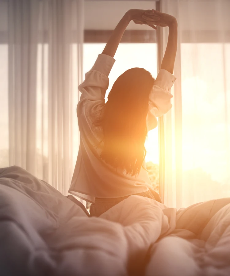 Woman stretching arms overhead in bed during peaceful morning routine in golden sunlight