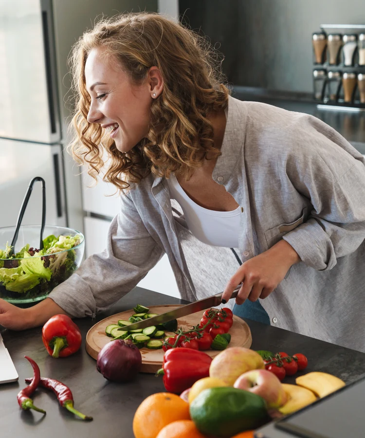 Happy woman preparing healthy salad with fresh vegetables while using laptop in modern kitchen
