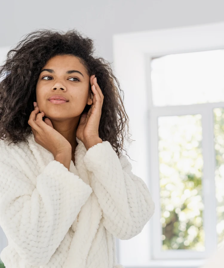 Woman in white robe touching face during skincare routine in bright bathroom mirror