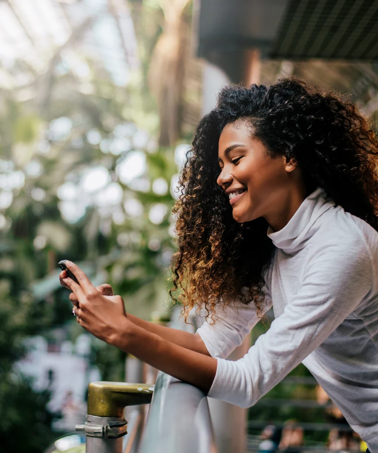 Happy woman with curly hair using smartphone for telehealth consultation in bright natural setting with plants