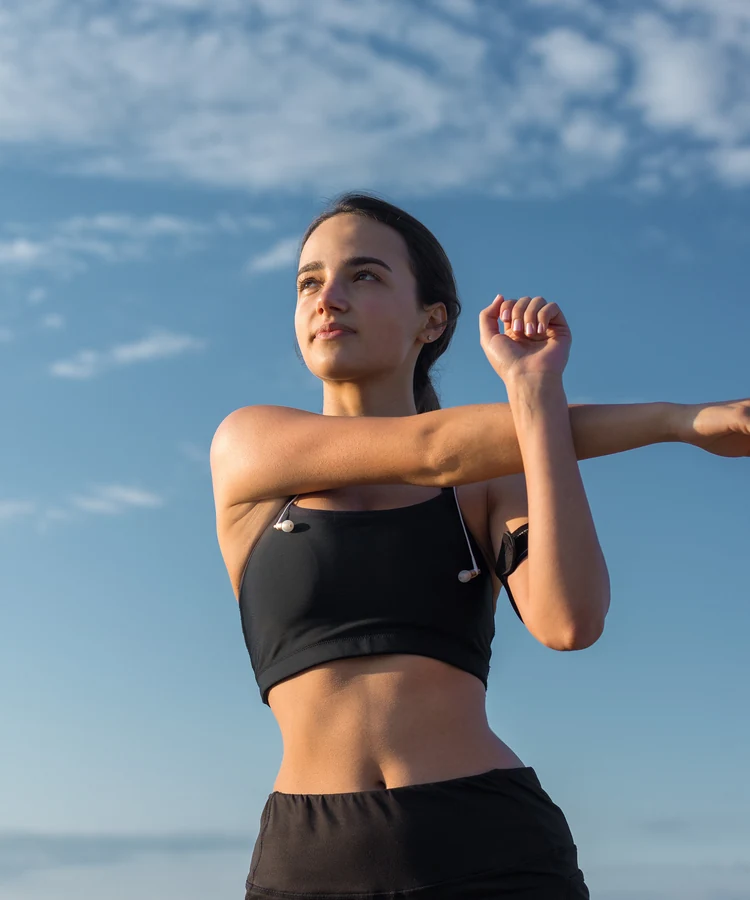 Confident woman in black athletic wear stretching arms outdoors against blue sky for fitness wellness routine