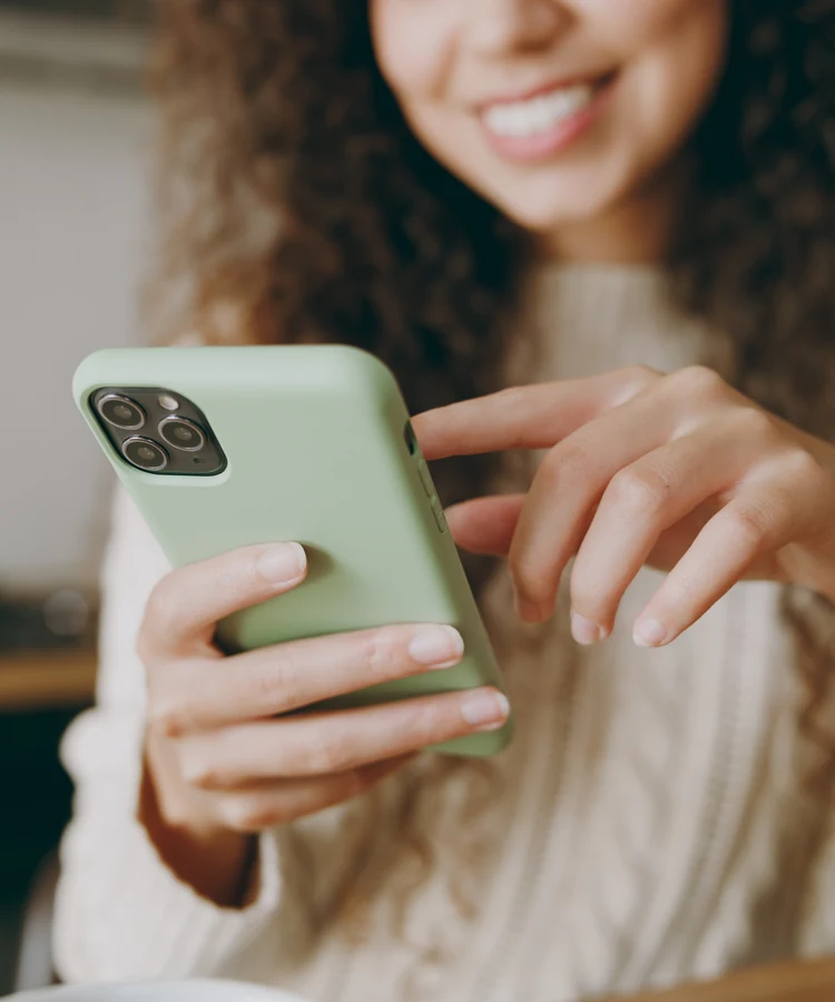Happy woman with curly hair smiling while using smartphone with green case in modern kitchen